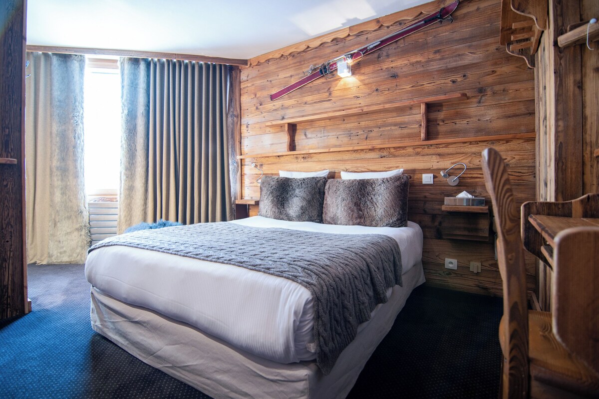 Une chambre d’hôtel avec une tête de lit en bois, un lit avec un matelas blanc et une couverture grise, une table de chevet en bois et un porte-skis fixé au mur.