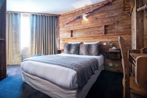 A hotel room with a wooden headboard, a bed with a white mattress and grey blanket, a wooden nightstand, and a ski rack on the wall.