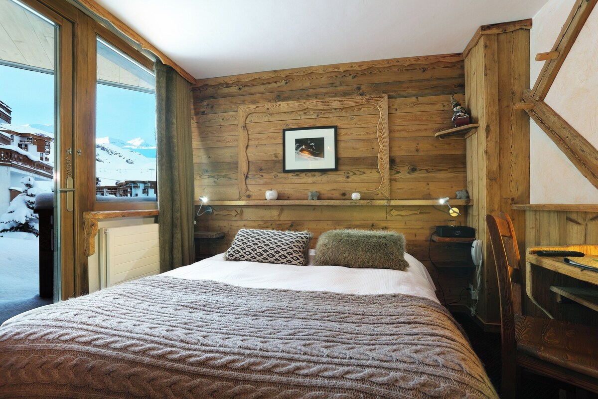 Une pièce chaleureuse aux murs en bois, avec un lit, une tête de lit en bois, un tableau encadré au mur et une vue sur un paysage enneigé par une grande fenêtre.