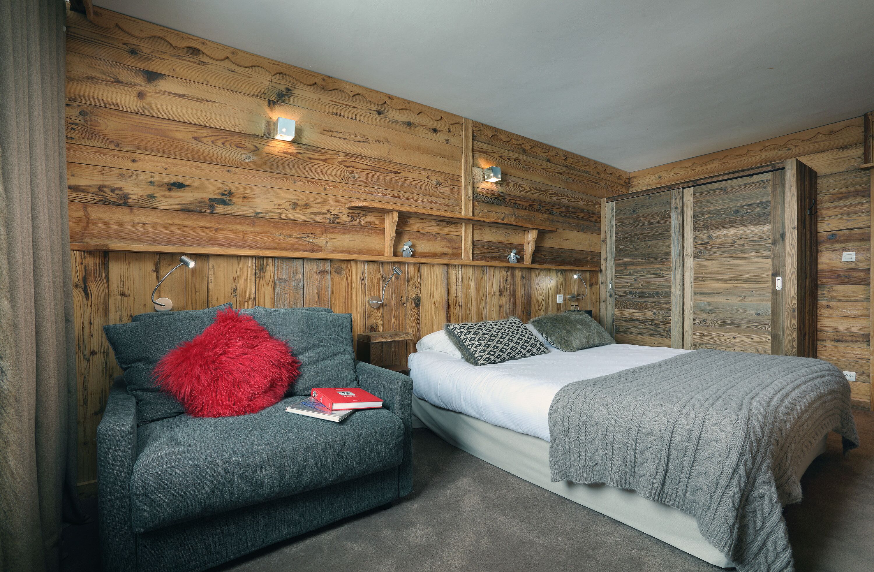 Um quarto com painel de parede de madeira, uma cama com manta cinza, um sofá cinza com almofada vermelha e uma mesa de cabeceira com abajur.