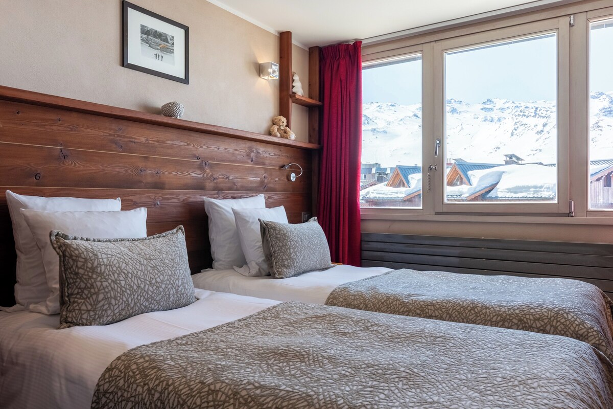 Une chambre d’hôtel avec deux lits, une tête de lit en bois, une fenêtre donnant sur des montagnes enneigées et un rideau rouge.