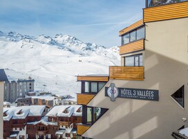 A hotel with a sign reading "Hôtel 3 Vallées" and a view of snow-covered mountains in the background.