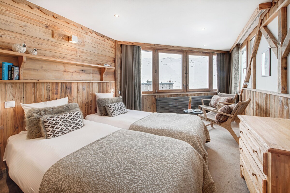 Une chambre confortable avec deux lits, une commode en bois, une chaise et une fenêtre donnant sur la montagne.