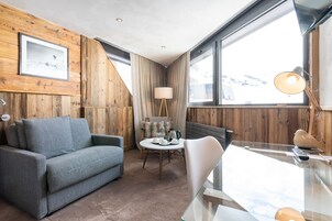 A cozy room with a wooden wall, a grey sofa, a round table with a lamp, and a view of mountains through large windows.