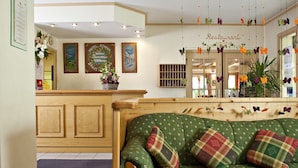 Un vestíbulo de hotel con mostrador de recepción, un sofá verde con cojines estampados, una mesa de centro de madera con una taza y un libro, y decoraciones de mariposas colgantes.