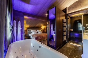 Une chambre d’hôtel moderne dotée d’une grande baignoire, d’un lit recouvert d’une couverture à carreaux et d’une salle de bain séparée avec une douche à parois de verre.