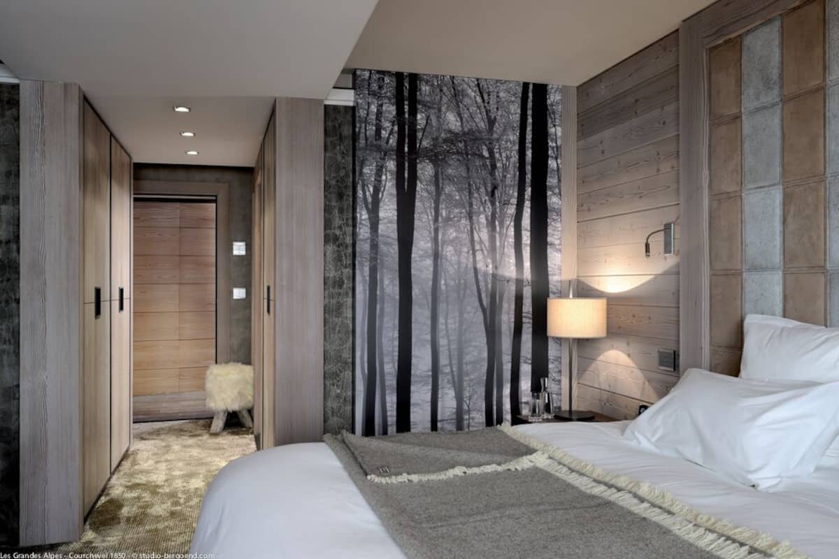 A modern bedroom with a large bed, a nightstand with a lamp, a wardrobe, and a forest-themed wall mural.