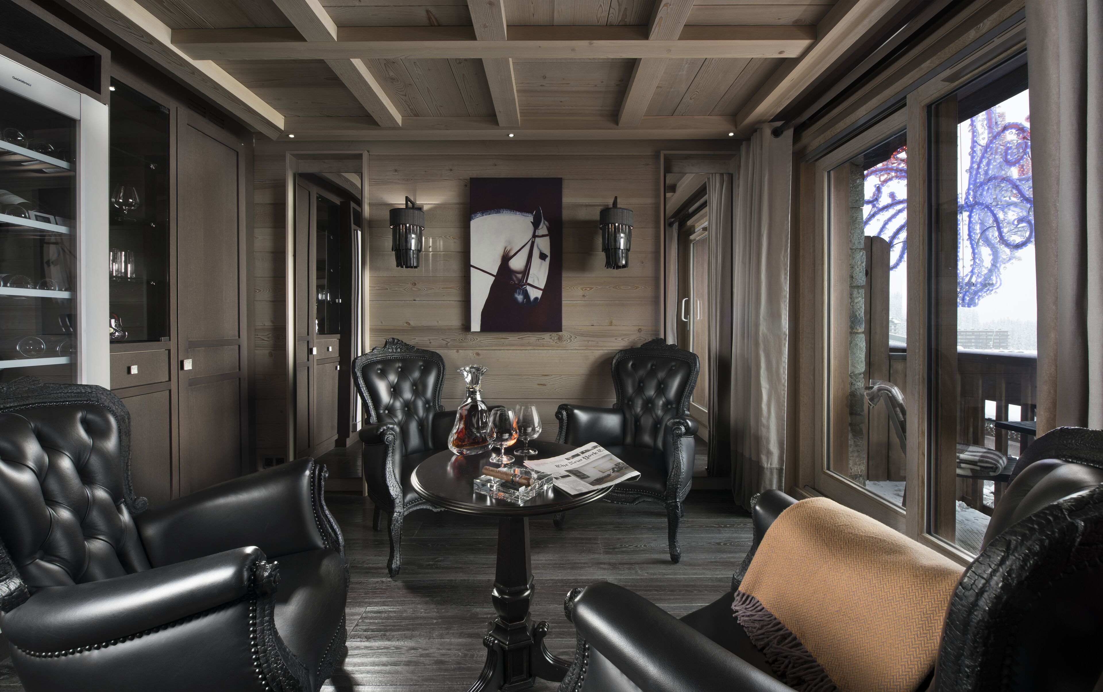 Une pièce avec un plafond en bois, une vitrine contenant des verres à vin, deux fauteuils en cuir, une table avec un journal et un tableau représentant la tête d’un cheval.