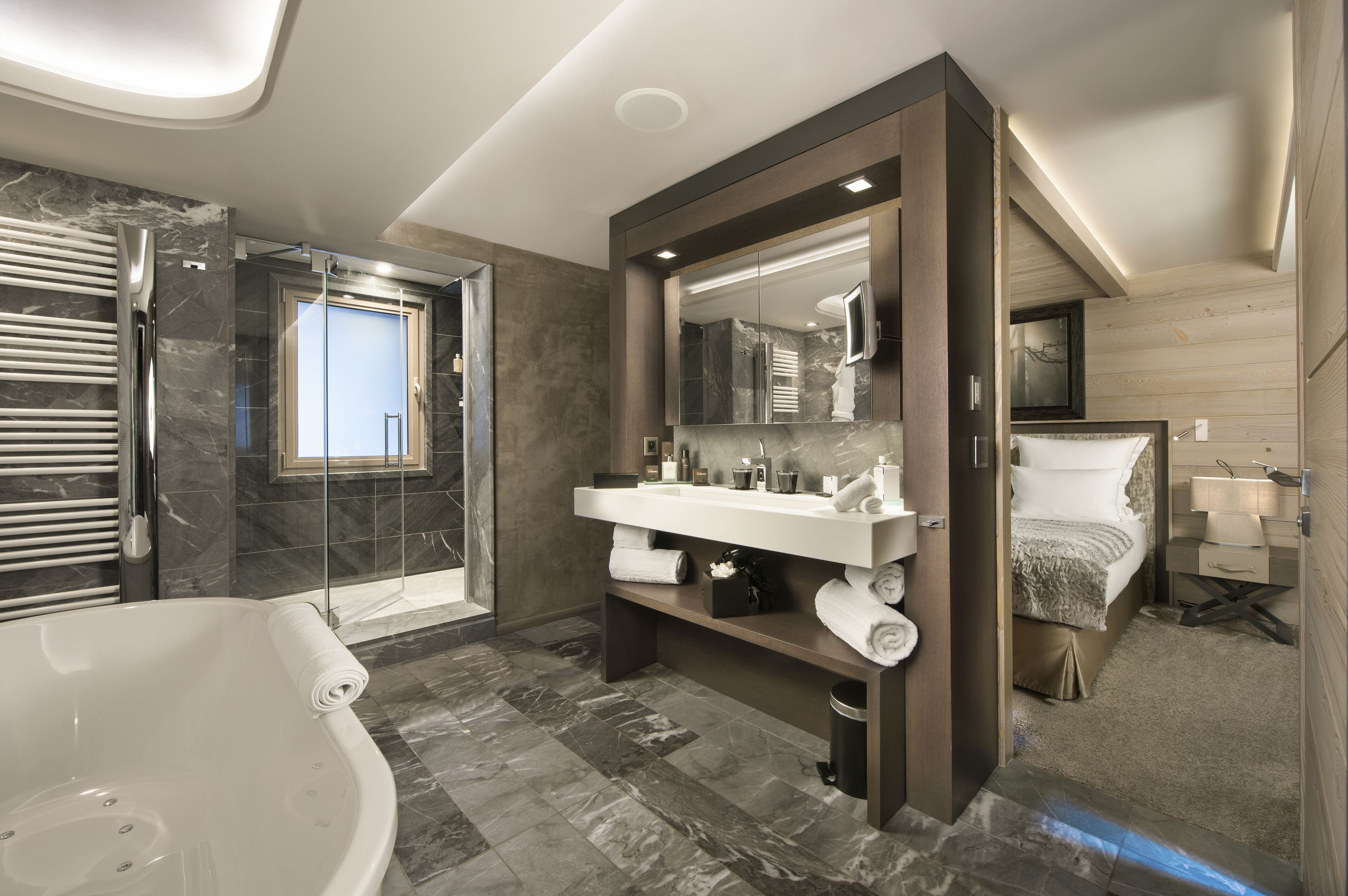 A modern bathroom with a large bathtub, a walk-in shower, a vanity with a mirror, and a bed in the corner.
