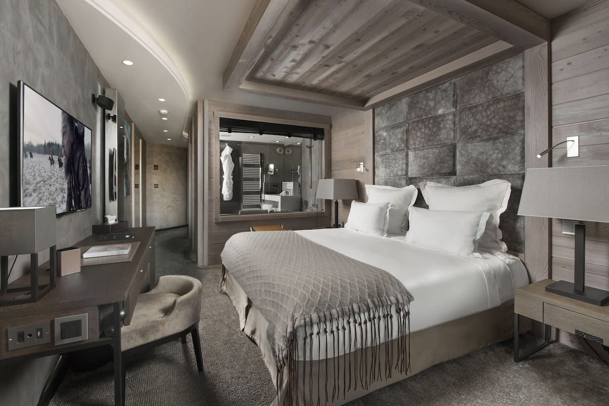 A modern hotel room with a large bed, a desk, a chair, and a bathroom visible through a glass door.