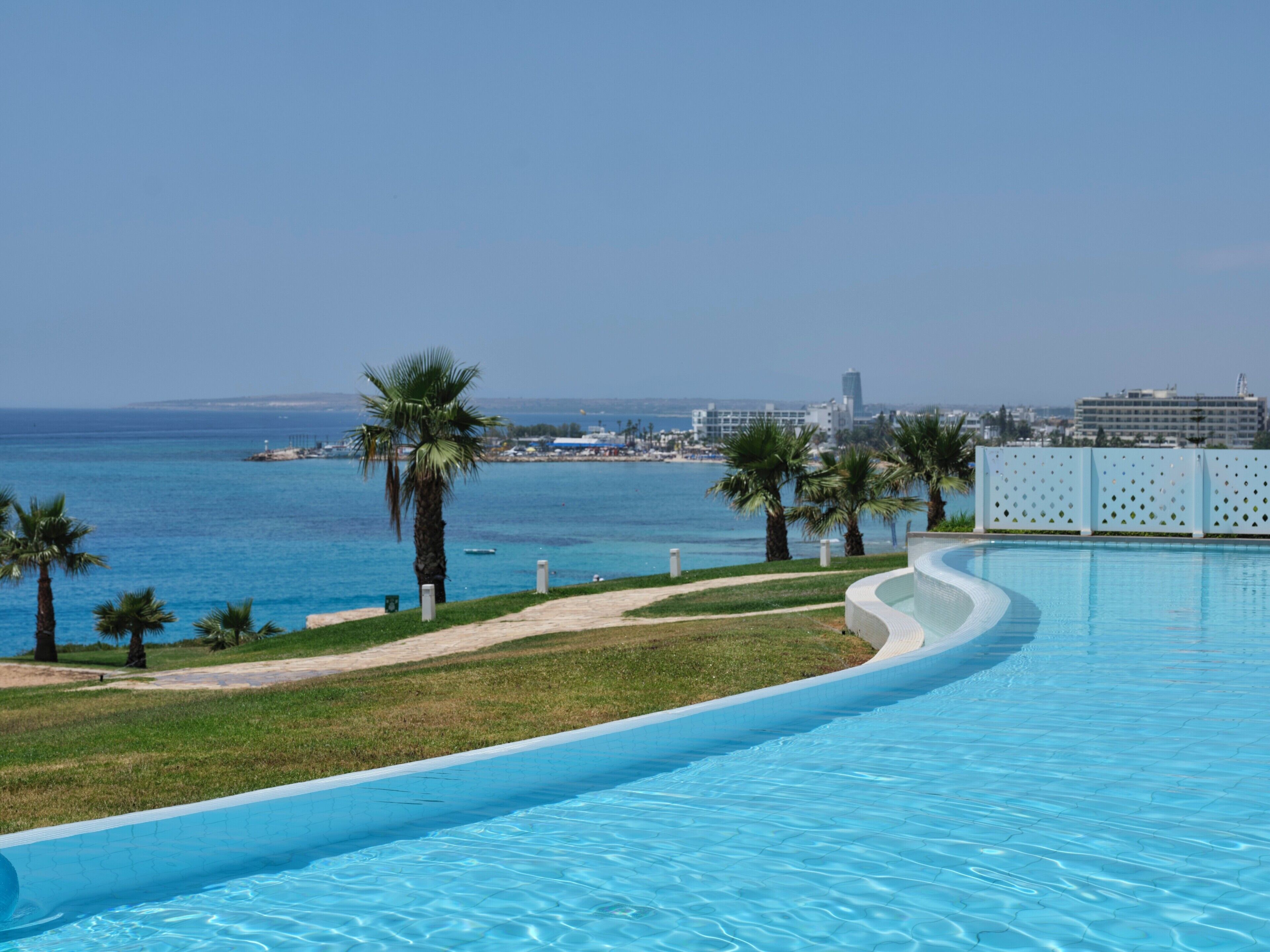Une piscine à bord incurvé, des palmiers et une vue sur la mer et les bâtiments en arrière-plan.