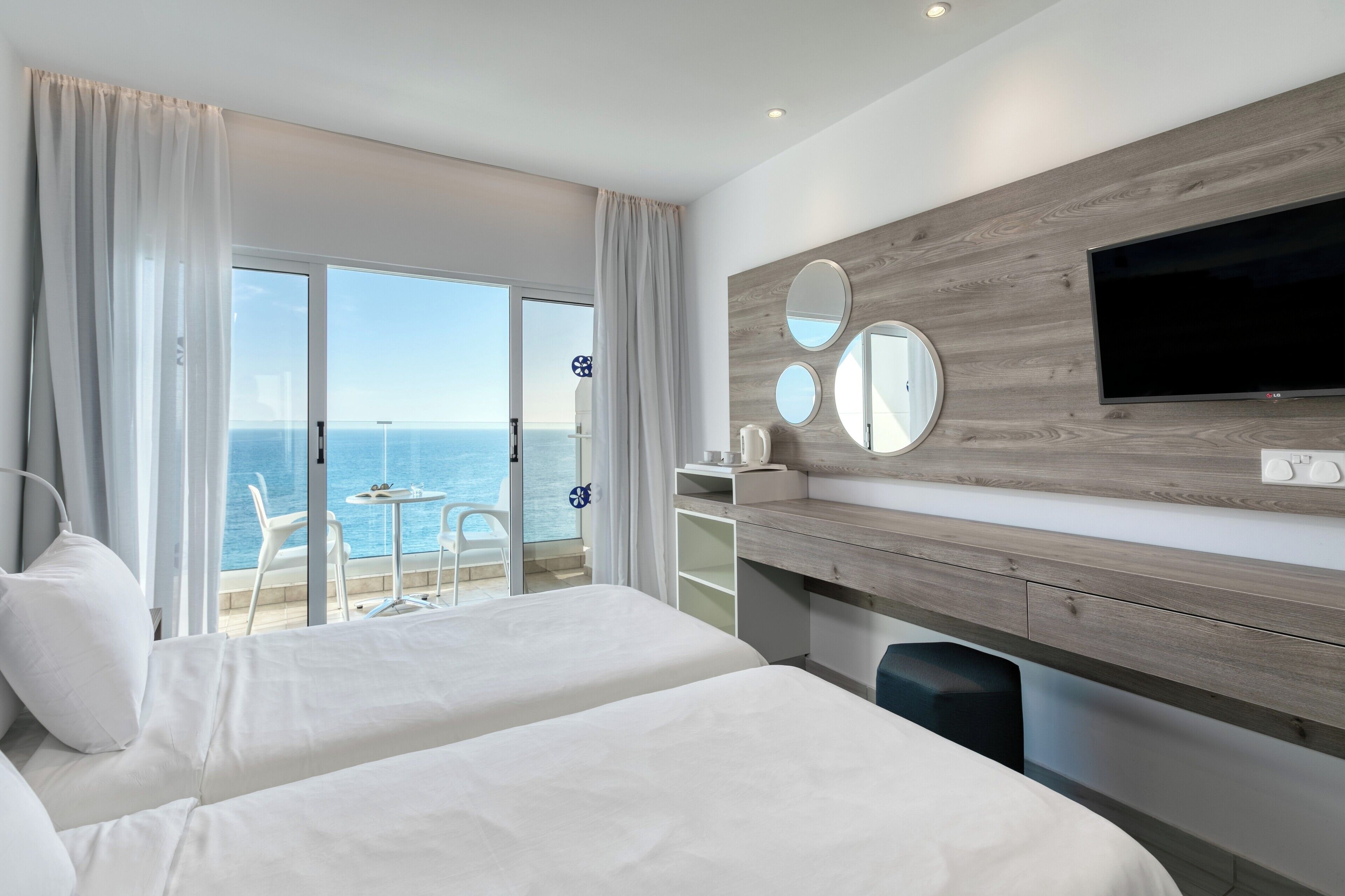 A hotel room with two beds, a TV, a desk, and a view of the ocean.
