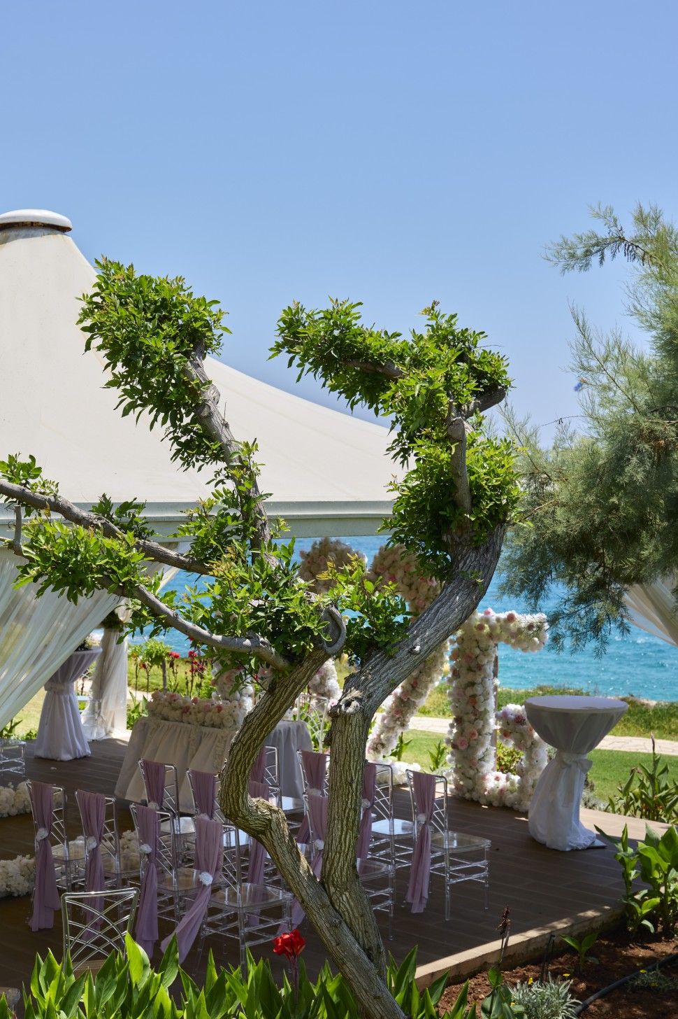 An outdoor wedding venue with a clear view of the sea, arranged tables with chairs, and a decorative archway.