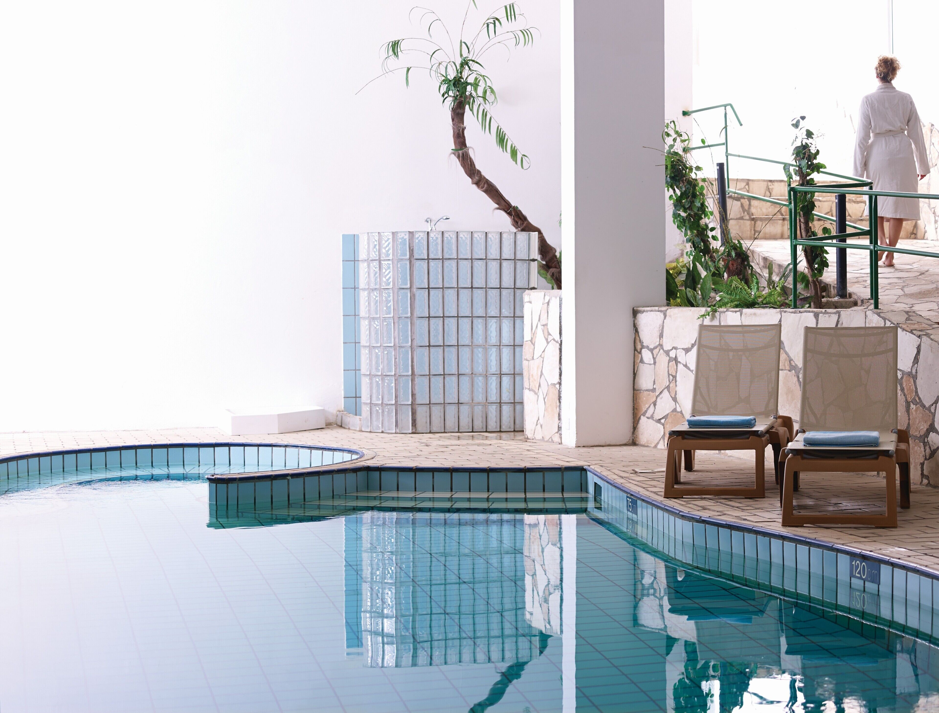 A swimming pool with a tiled edge, a stone wall, and a staircase leading to a balcony with plants.