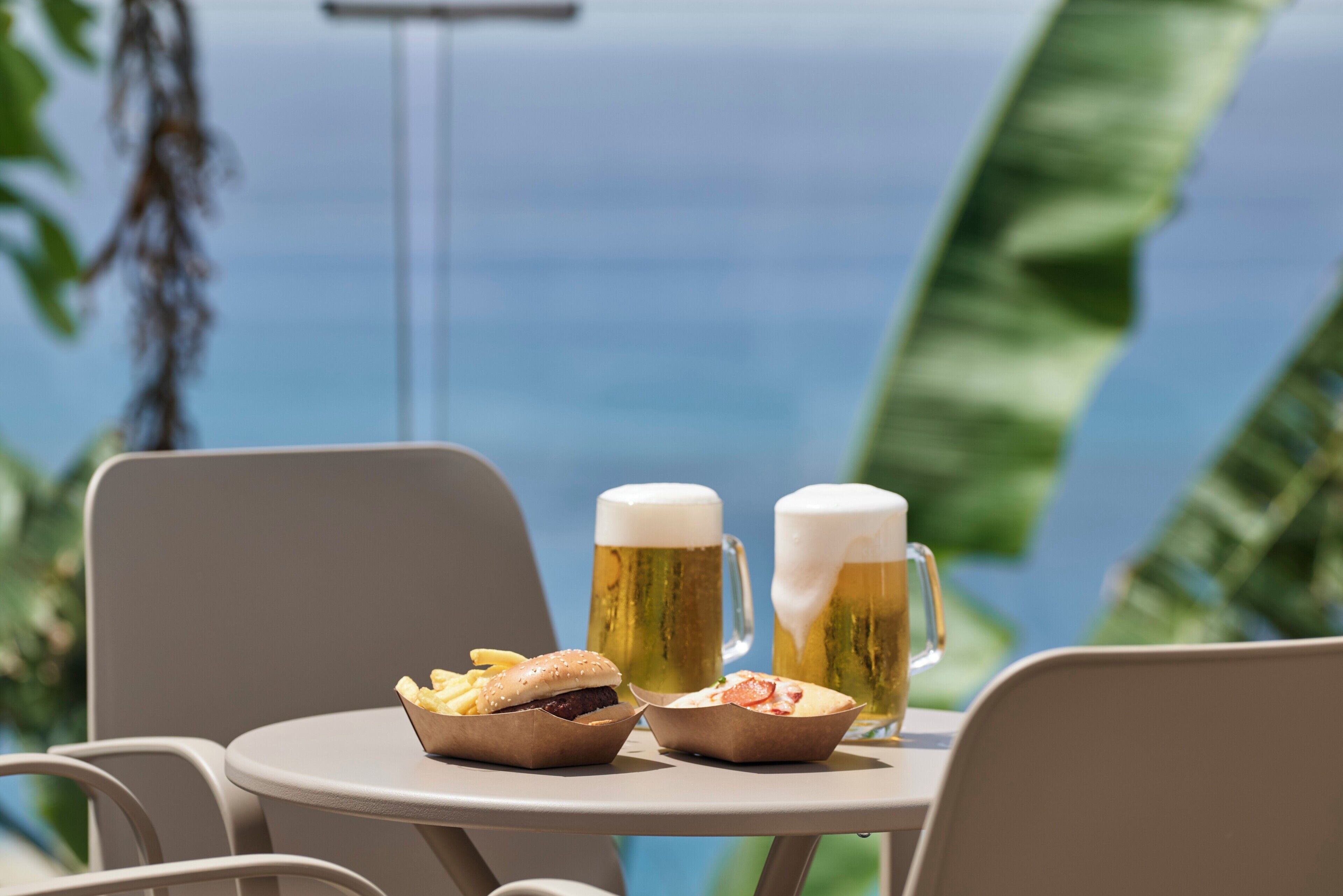 A table with two beer mugs and a burger, set against a backdrop of a clear sky and ocean view.
