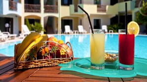 A poolside setting with a wicker fruit basket, two drinks with straws, and a pool in the background. - Ariadimari Hotel (Valledoria)