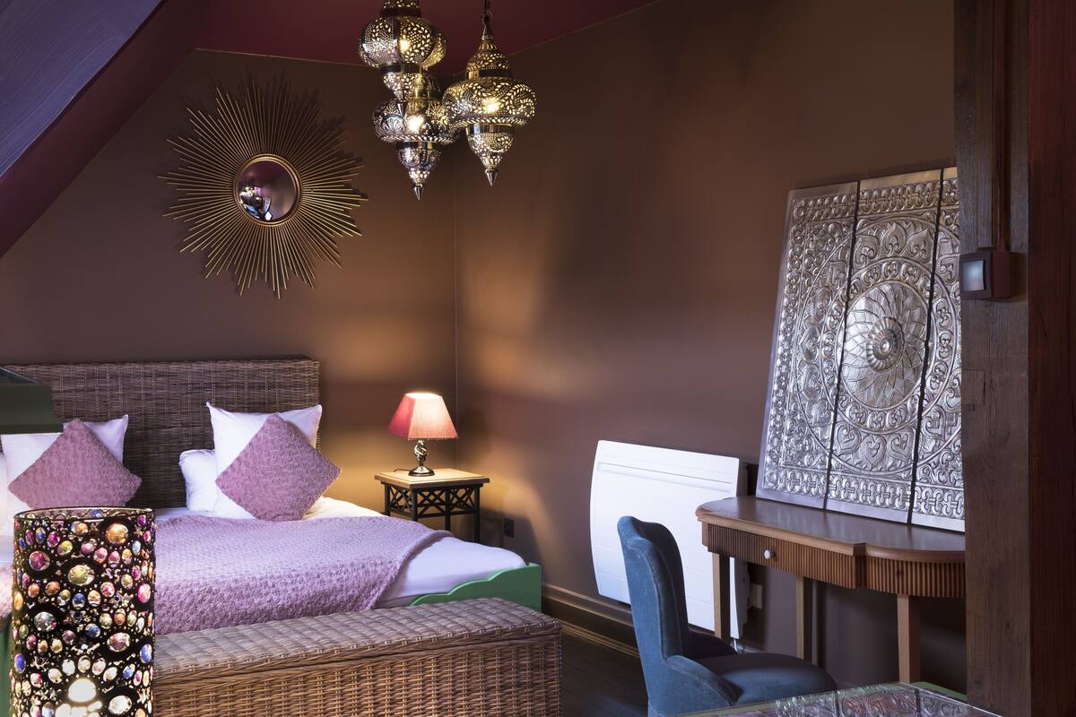 A bedroom with a bed, a nightstand, a lamp, a chair, a decorative wall piece, and a hanging light fixture.