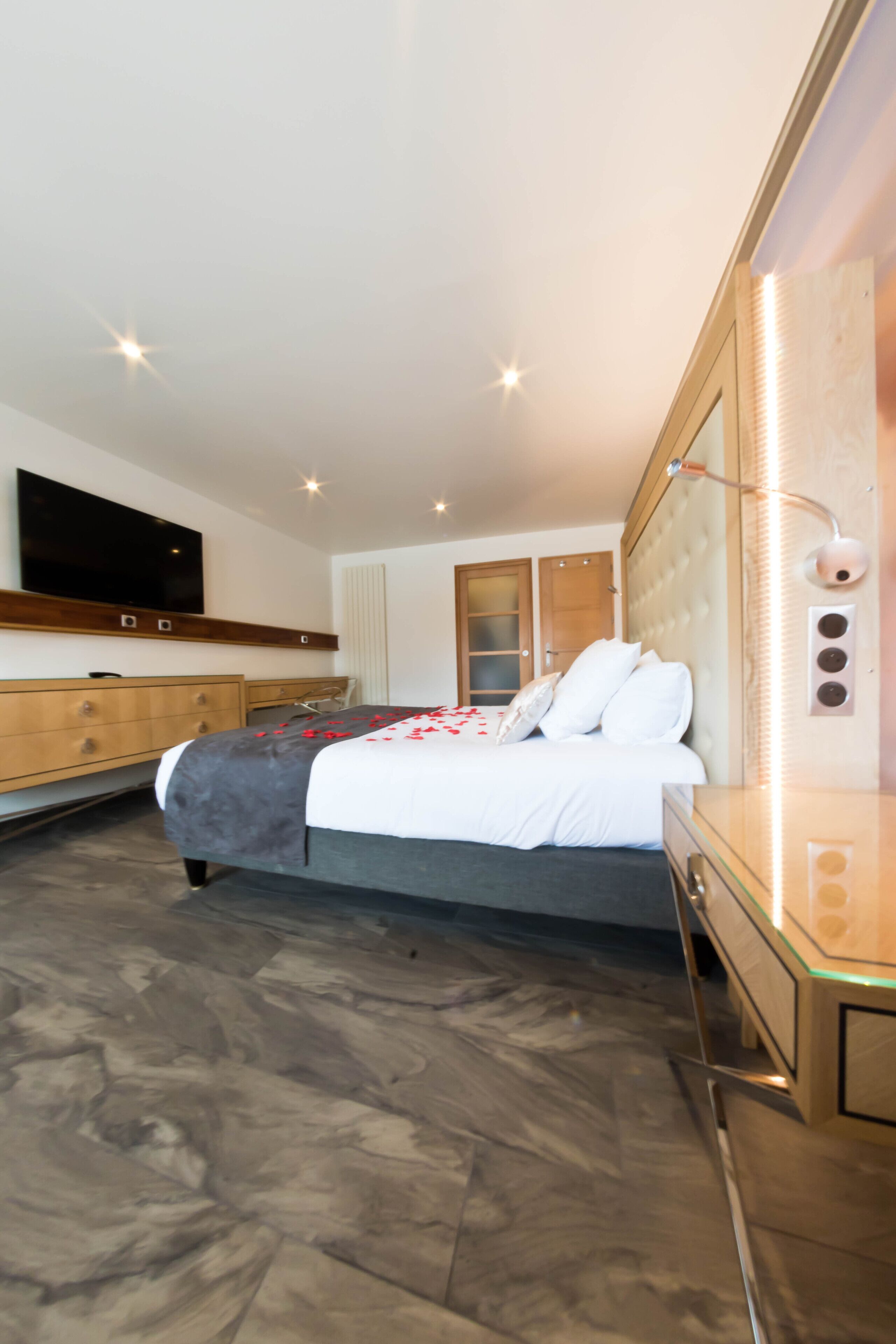A modern bedroom with a large bed, a TV mounted on the wall, a wooden dresser, and a glass cabinet.