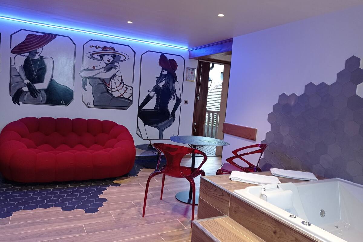 A modern bathroom with a wooden floor, a red bean bag chair, a glass bathtub, and hexagonal tiles.