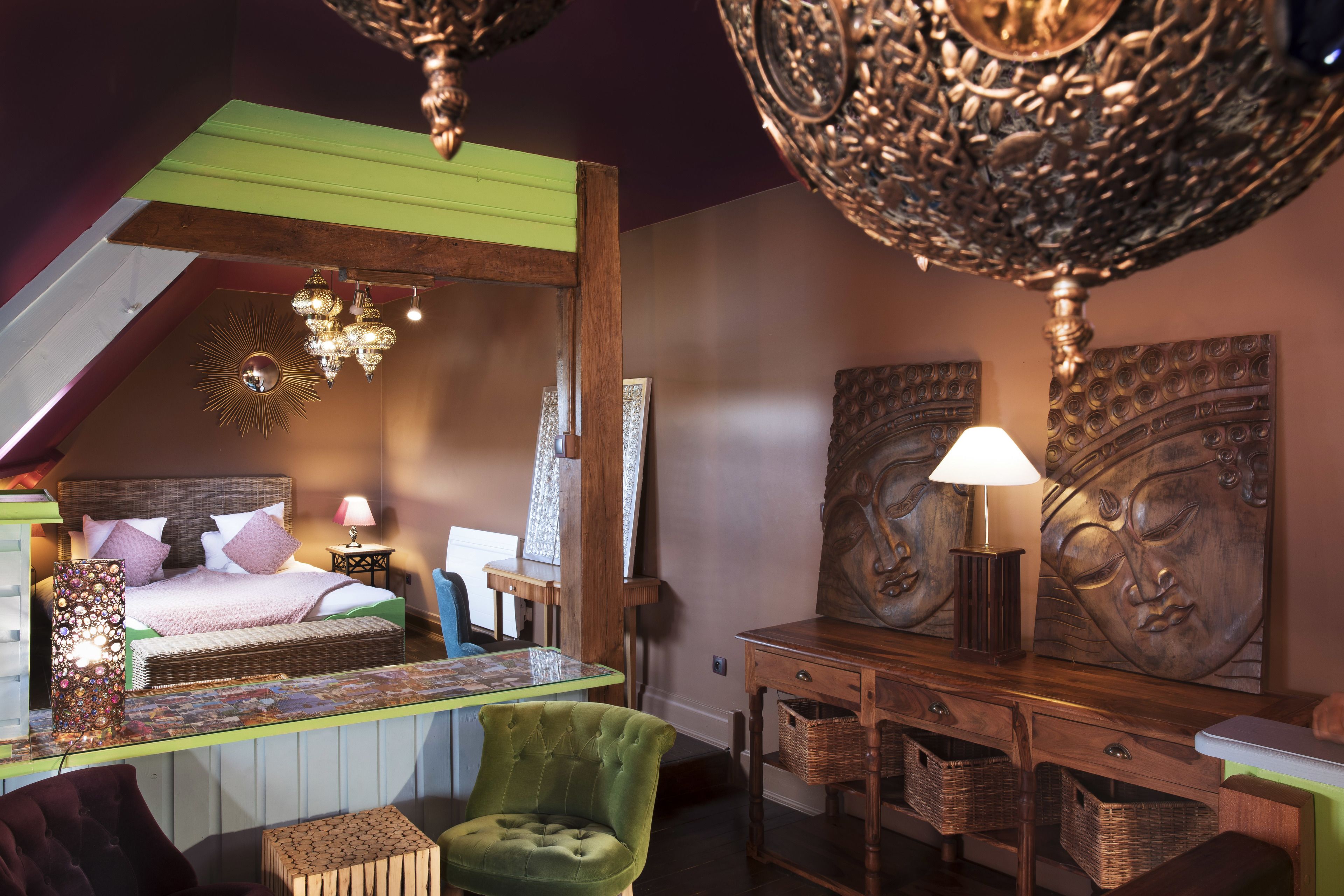 Une chambre avec un plafond en pente, un lit, un fauteuil vert, une commode en bois et un lustre.