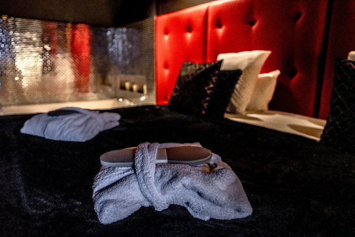 A hotel room with a red tufted headboard, a glass shower enclosure, and a black bath mat.