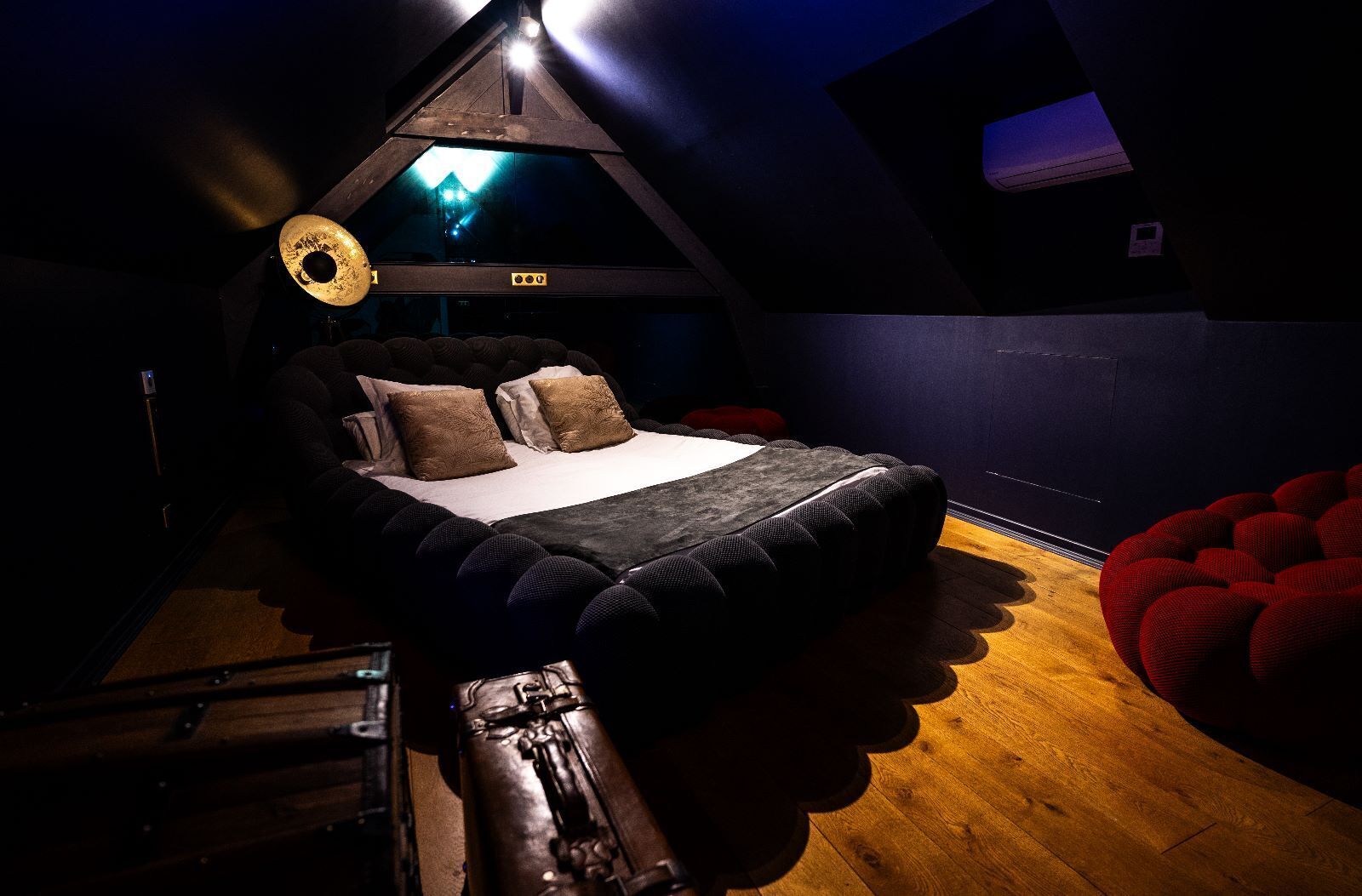 Ein Schlafzimmer im Loft-Stil mit schrägem Dach, einem Bett mit dunklem Kopfteil und einem roten Hocker.