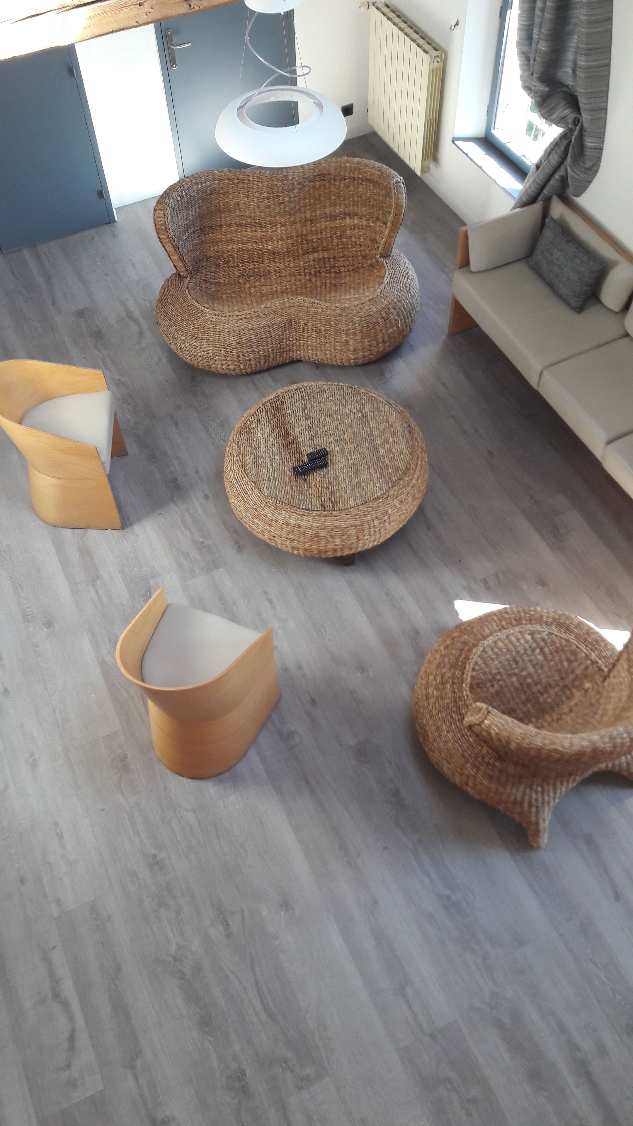 A room with a grey wooden floor, a woven round chair, a woven stool, a wooden chair, and a sofa with a cushion.