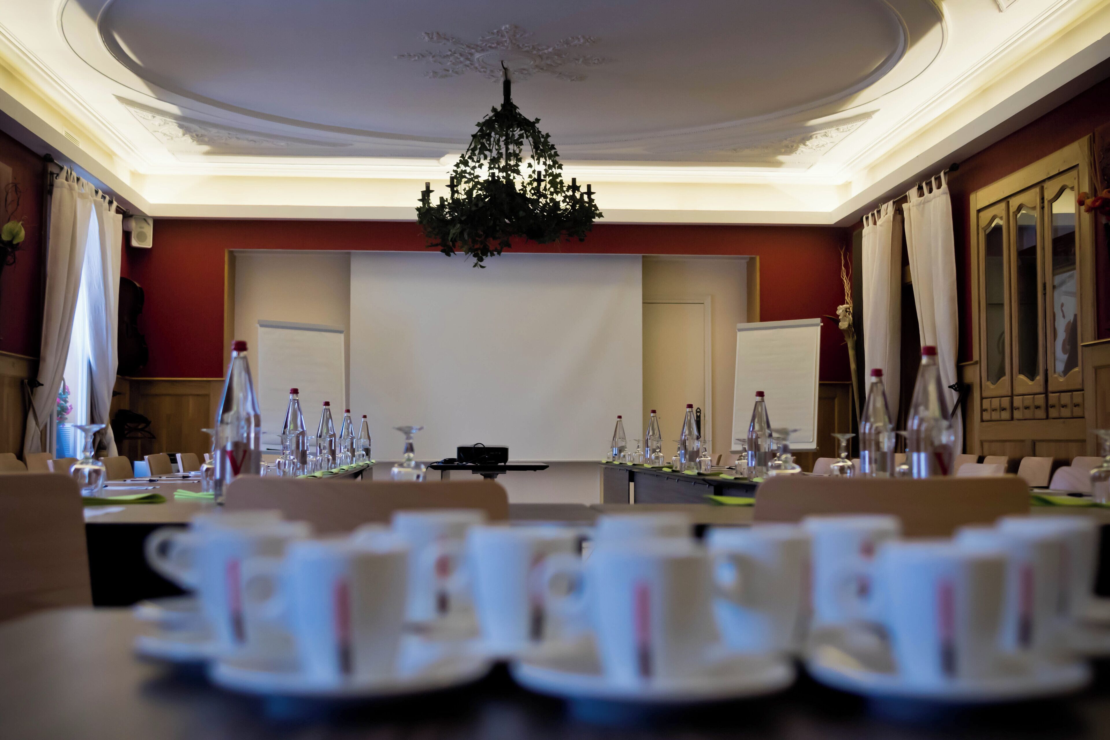 Un salón de conferencias con una gran mesa dispuesta para una reunión, una pantalla de proyector y una araña.