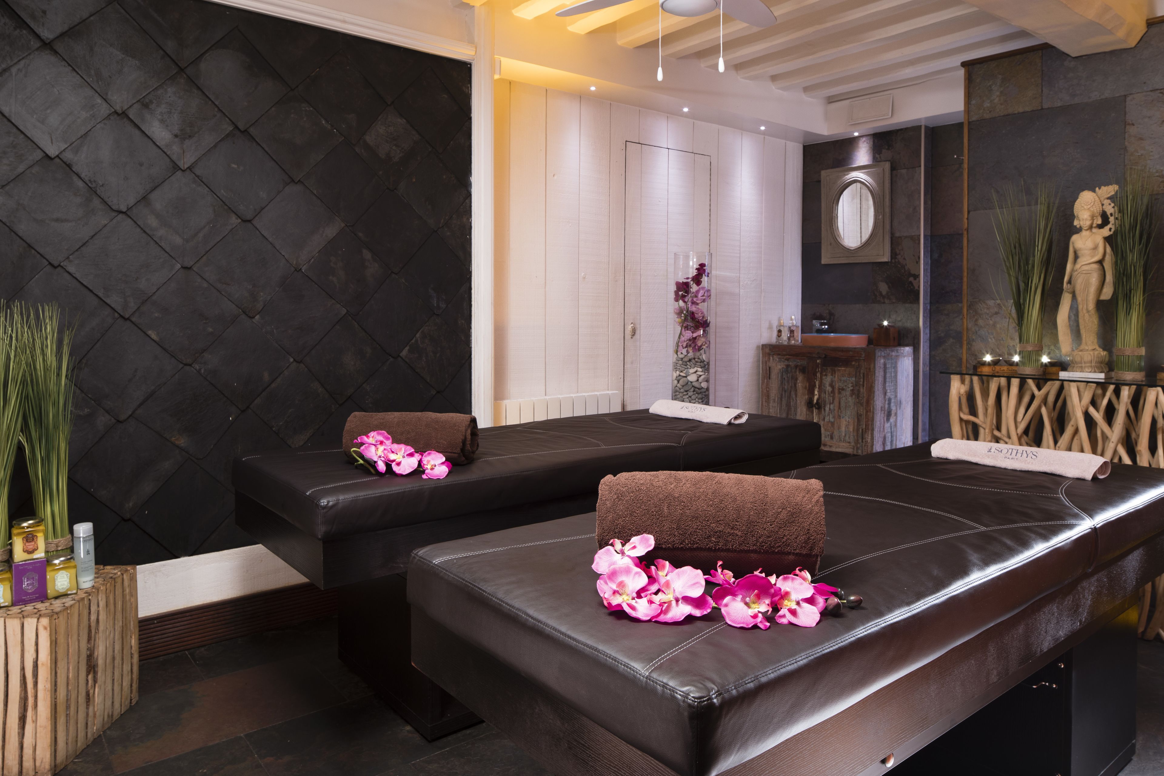 Un cuarto de spa con dos camillas de masaje, cada una con una toalla y flores rosadas. Hay una mesita de madera con botellas y un espejo en la pared.