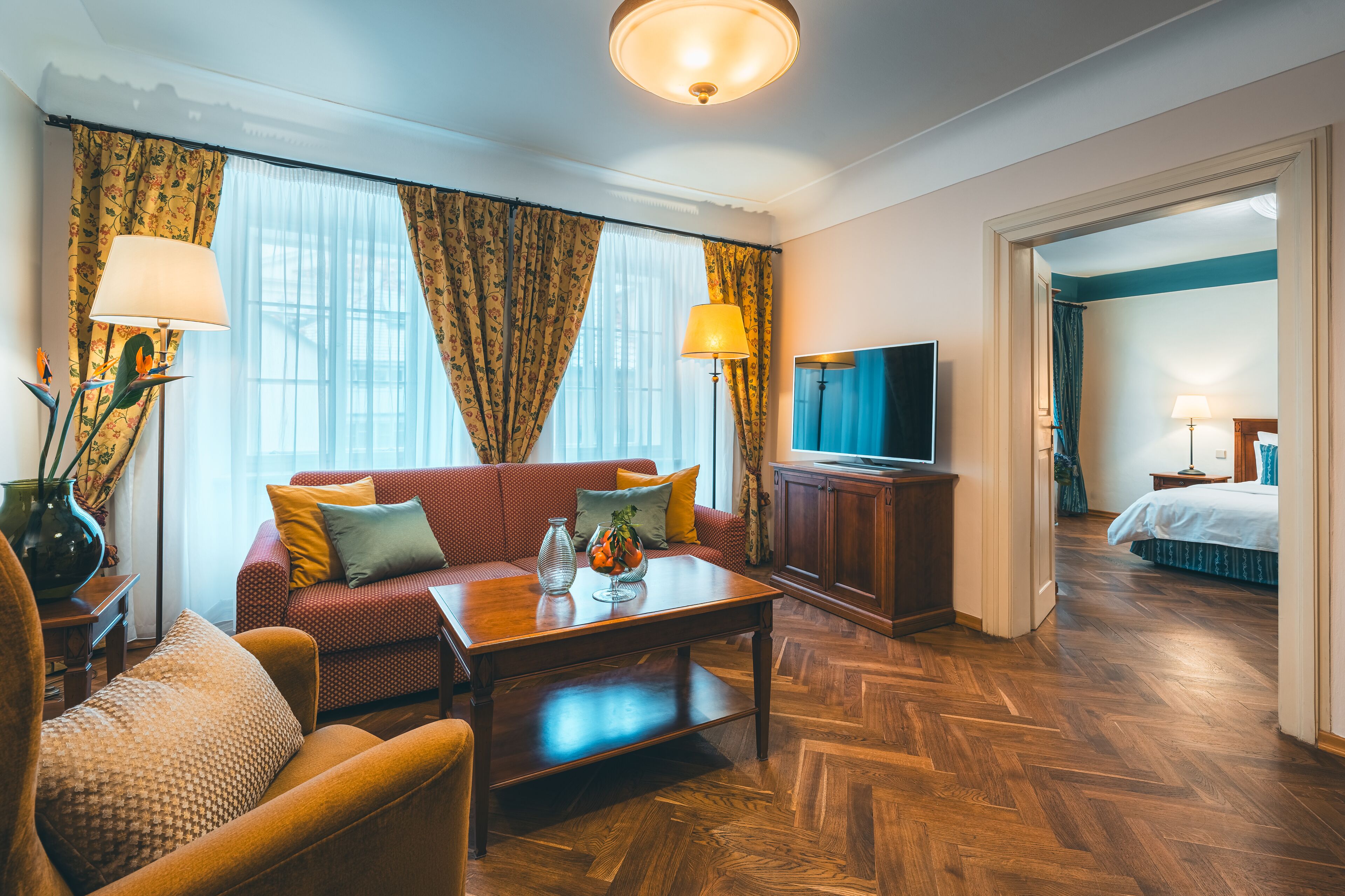 A hotel room with a sofa, armchair, wooden coffee table, TV, and a bed in the adjacent room.
