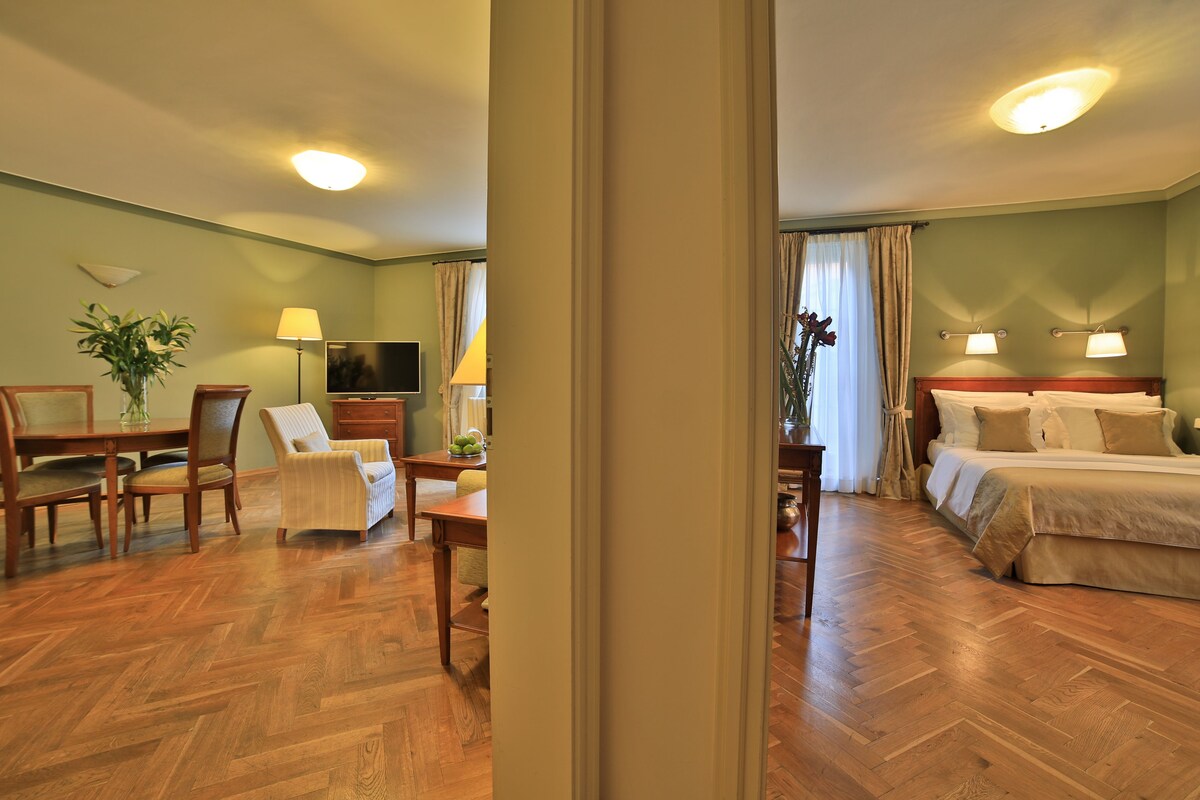 Habitación de hotel con suelo de madera en patrón de espiga, zona de comedor con mesa y sillas, televisor y cama con cabecero y almohadas.