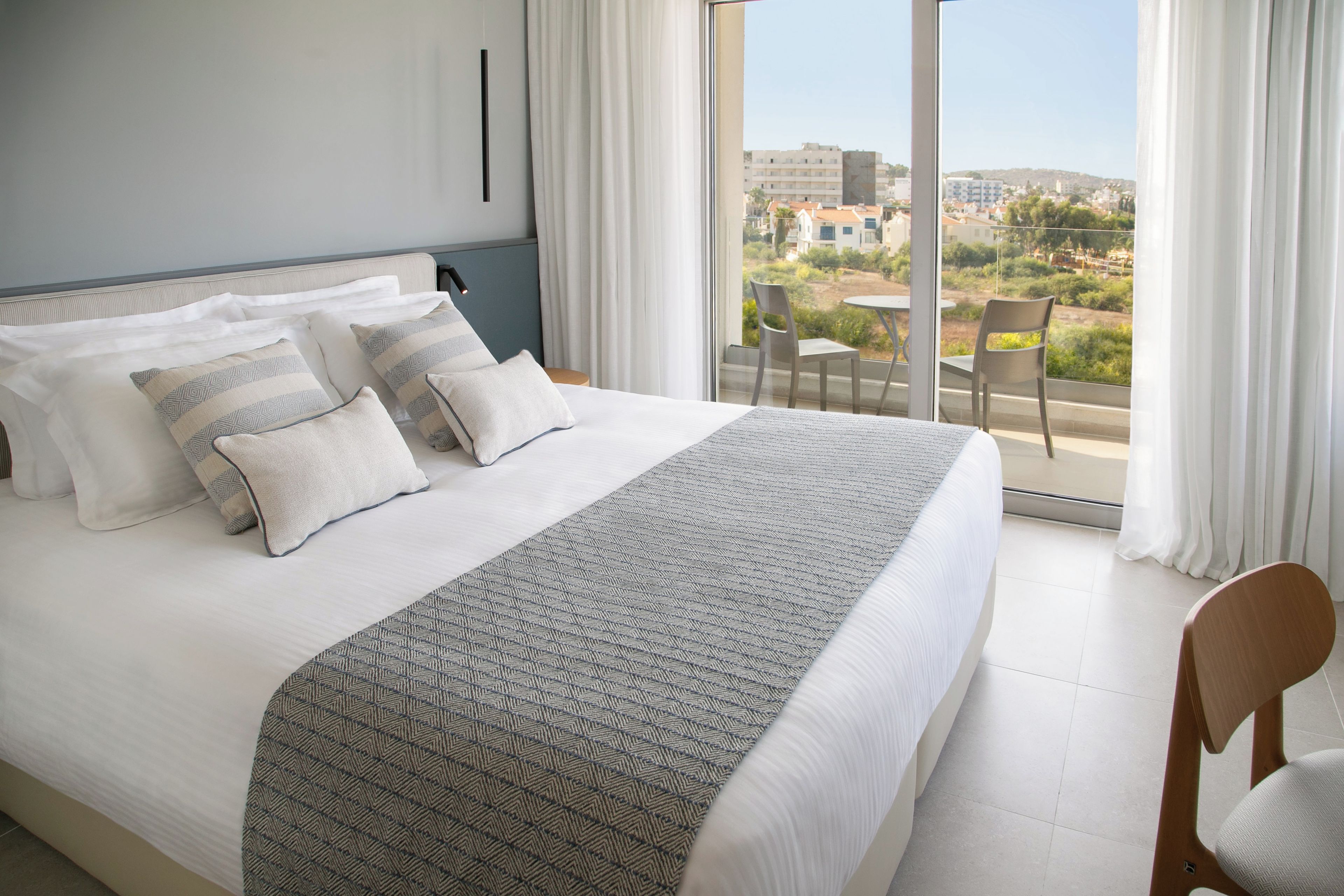 A hotel room with a large bed, a view of the city through the balcony, and a dining area with chairs.