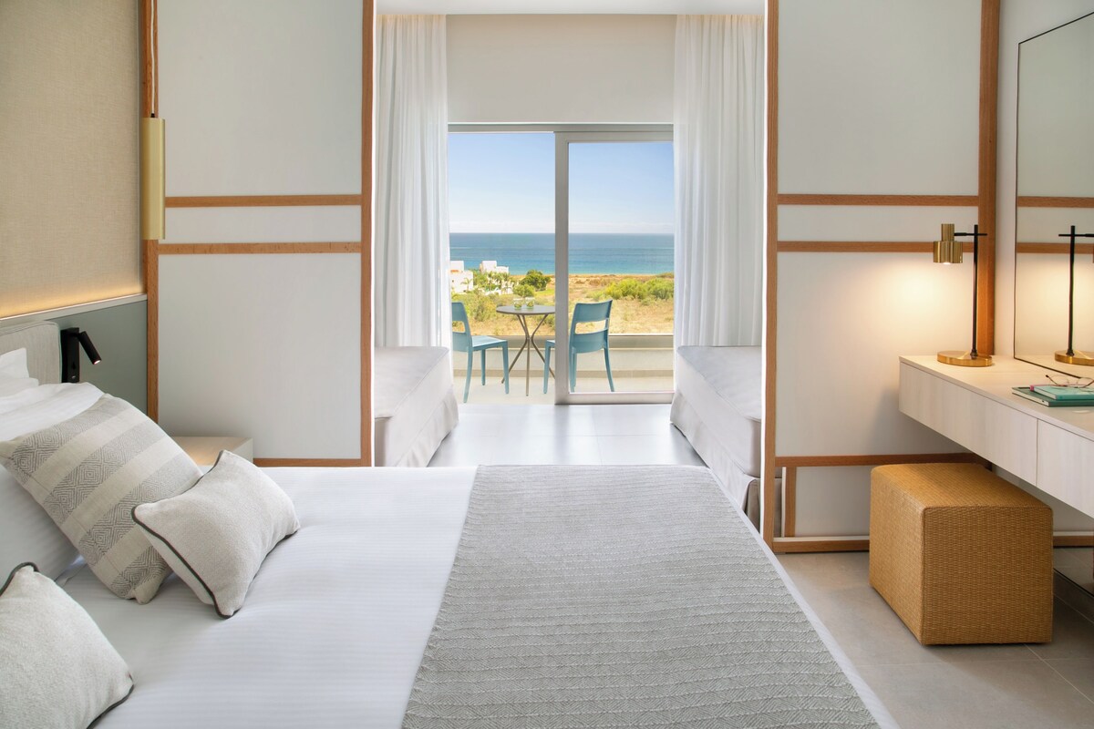 A hotel room with a bed, a desk, a chair, and a view of the sea.