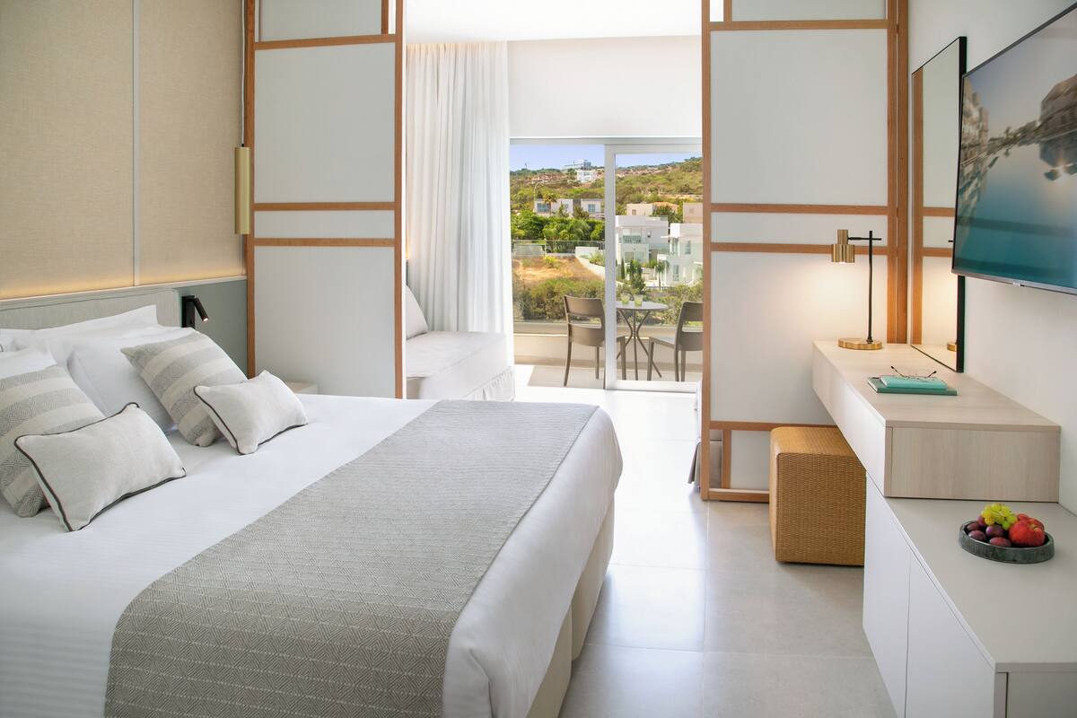 Quarto de hotel moderno com uma cama grande, uma escrivaninha com computador, televisão e varanda com vista para a paisagem.