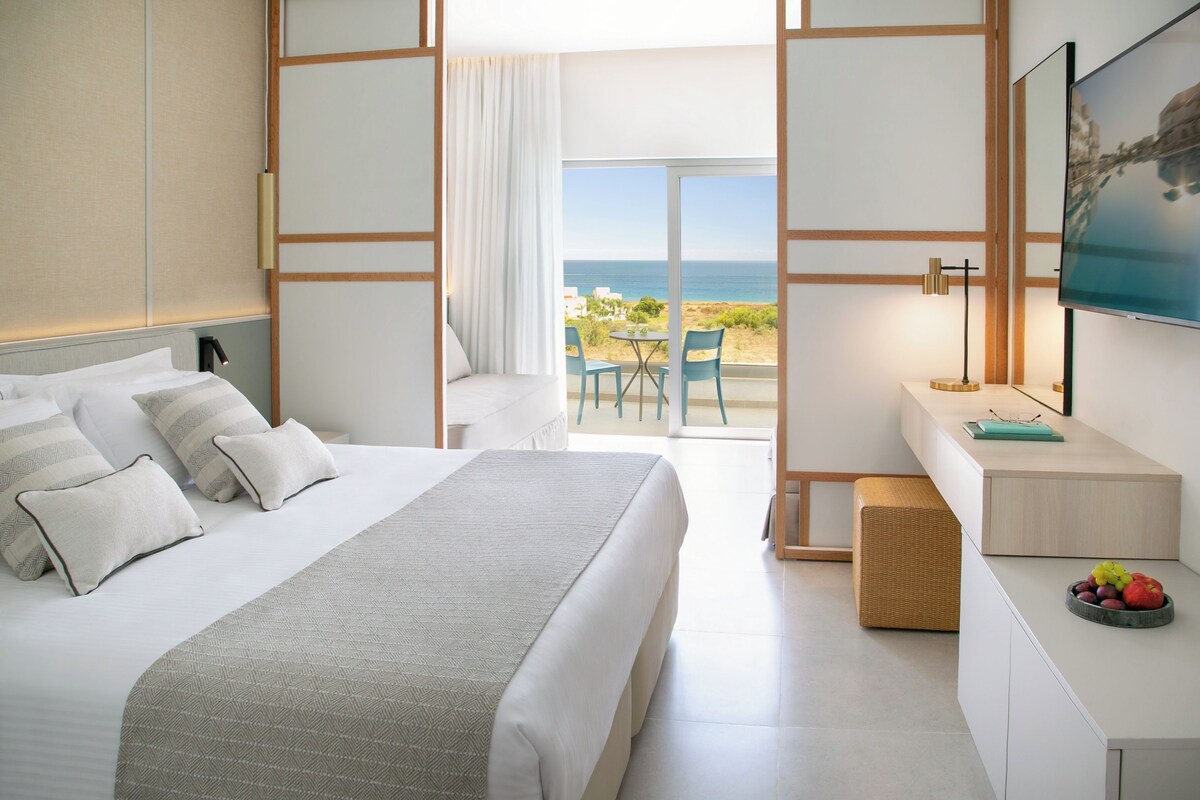 A modern hotel room with a large bed, a desk with a computer, a chair, and a view of the sea.