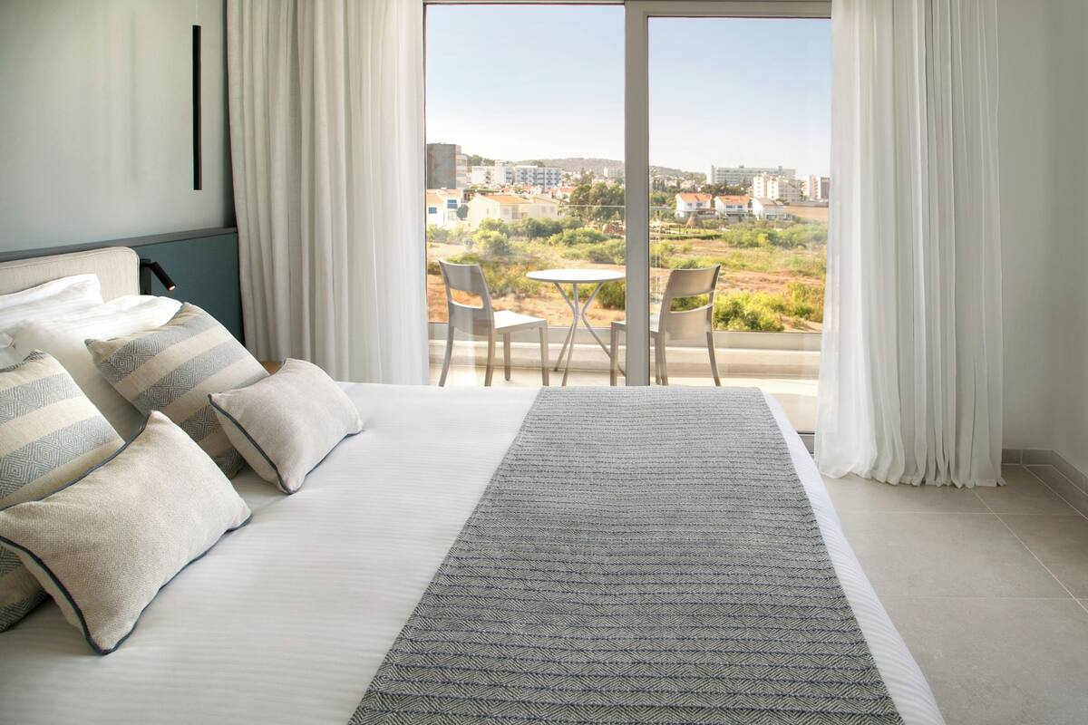 Quarto de hotel moderno com uma cama grande, roupa de cama branca, tapete cinza e uma varanda com vista para a cidade.