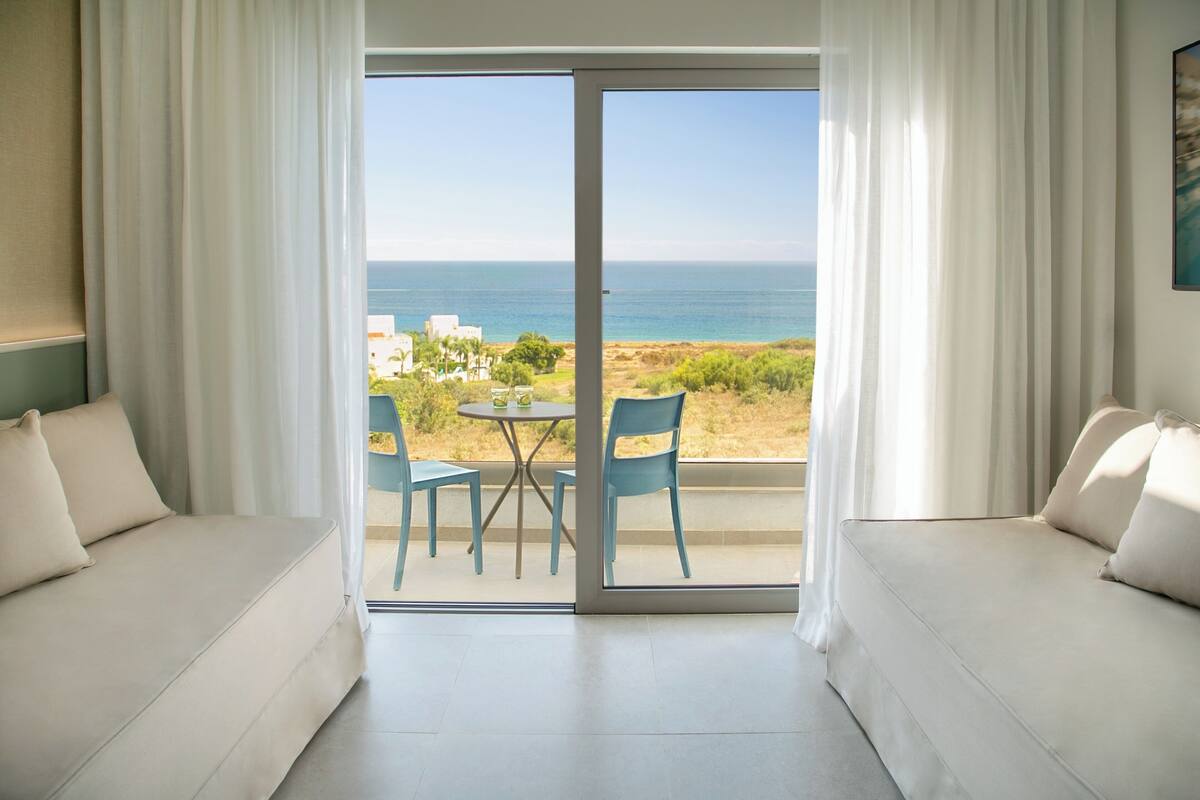 Quarto com sofá, mesa e cadeiras, e vista para o mar.