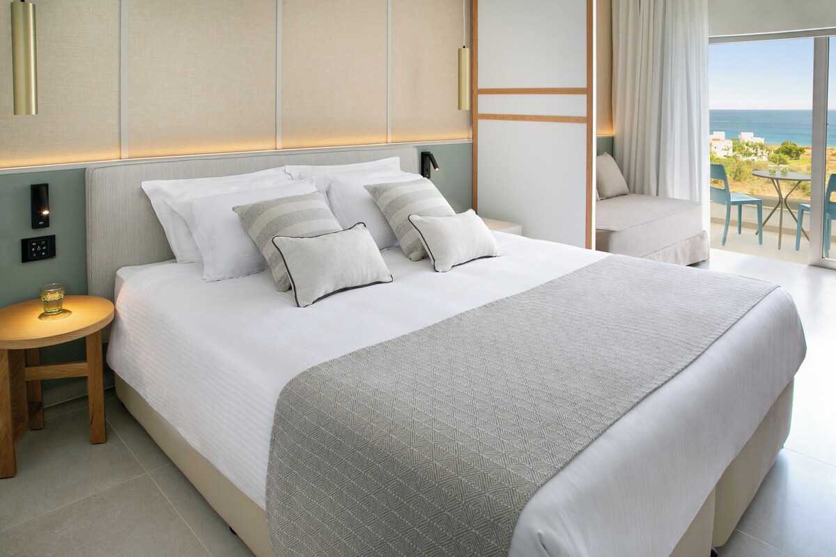 Une chambre d’hôtel moderne avec un grand lit, des commandes situées près du lit, une petite table avec une boisson, et un balcon avec vue.