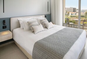 A neatly made bed with a patterned throw, a bedside table with a lamp, and a view of the city through large windows. - Atlantica Aqua Blue (Paralimni)