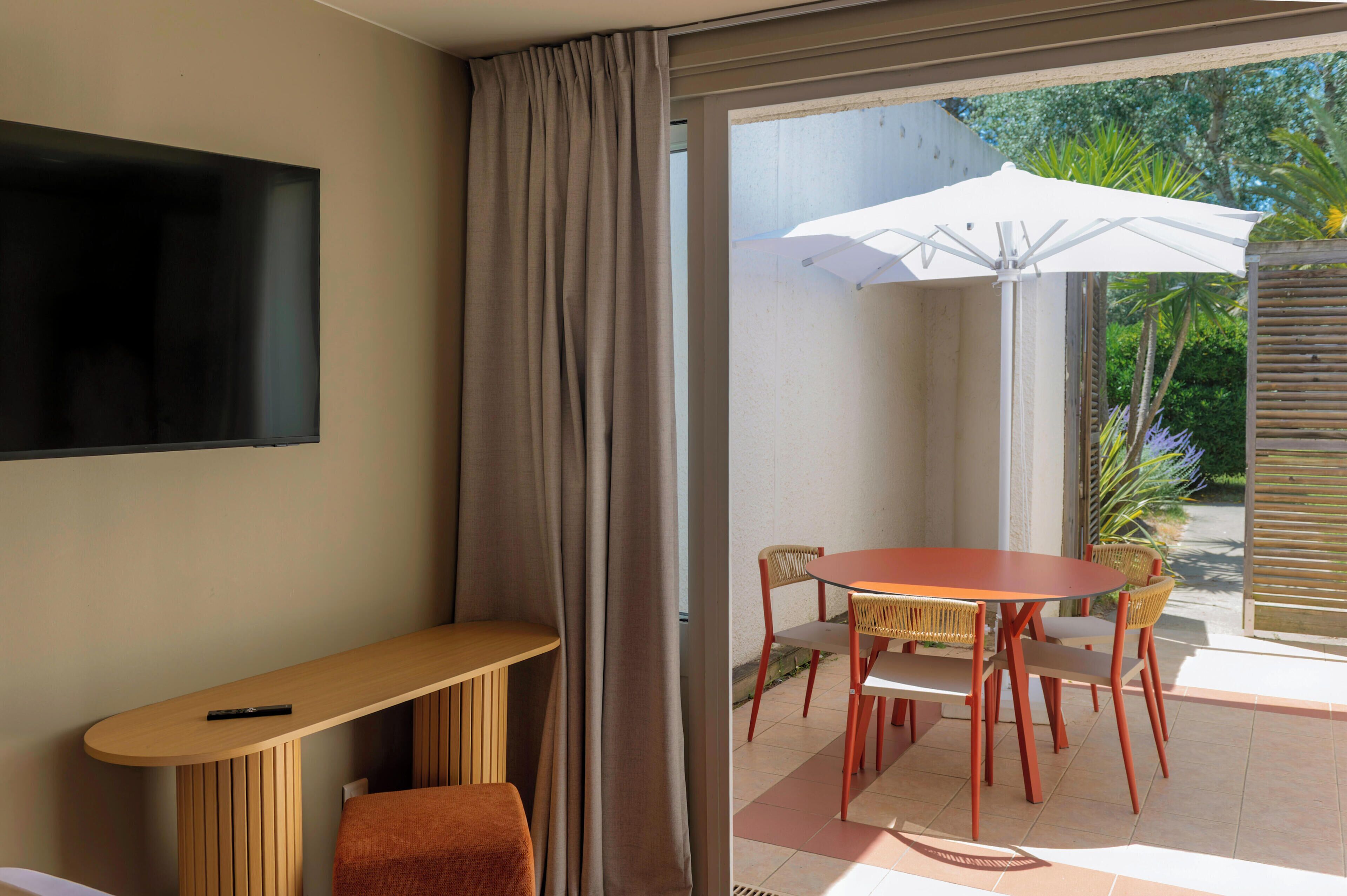 A room with a TV mounted on the wall, a wooden bench, a small table, and a dining area with chairs and an umbrella.