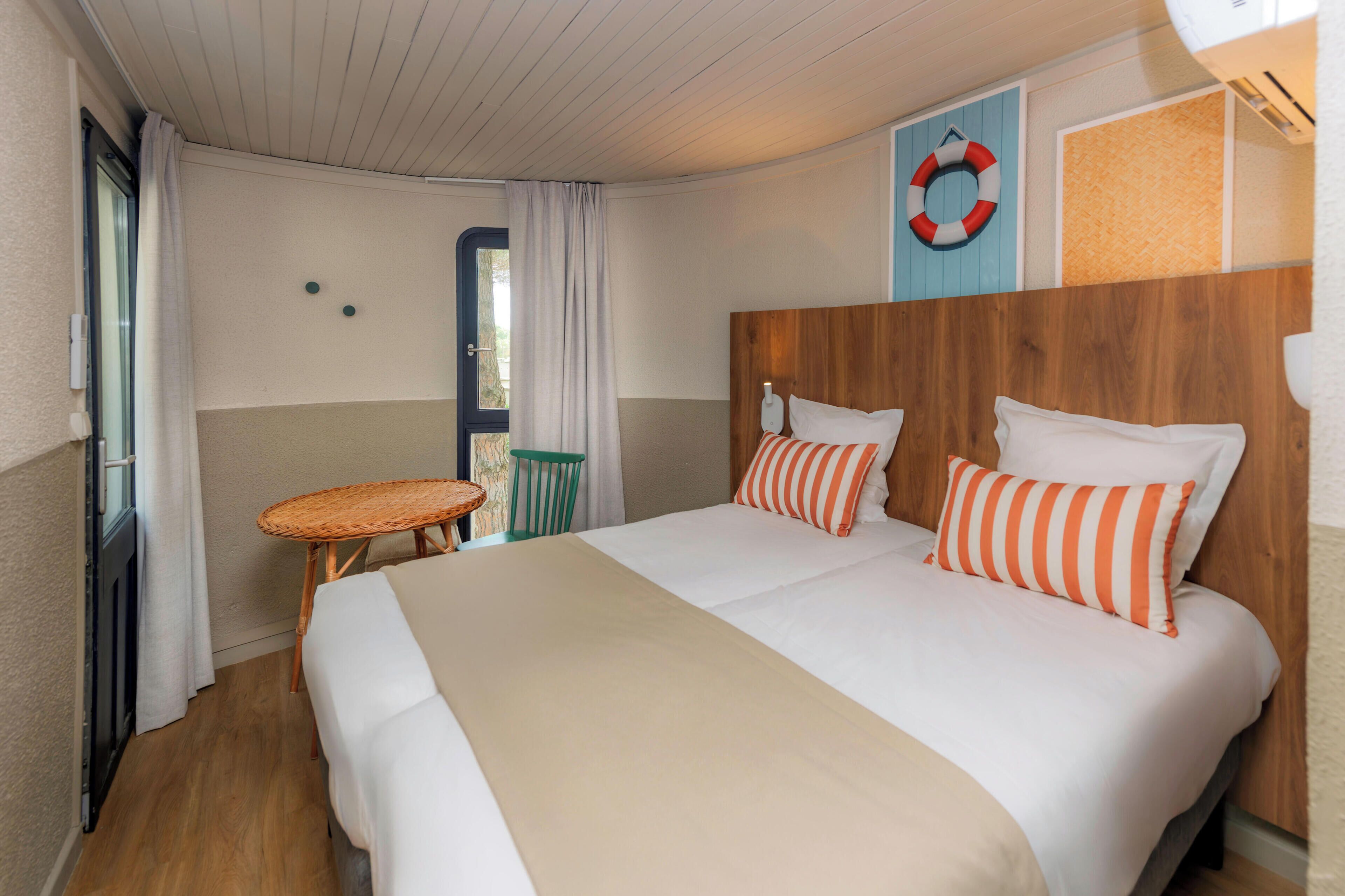 Ein Hotelzimmer mit Bett, einem kleinen Tisch, einem Stuhl, einem Fenster mit Vorhängen und einer Rettungsboje als Wanddeko.