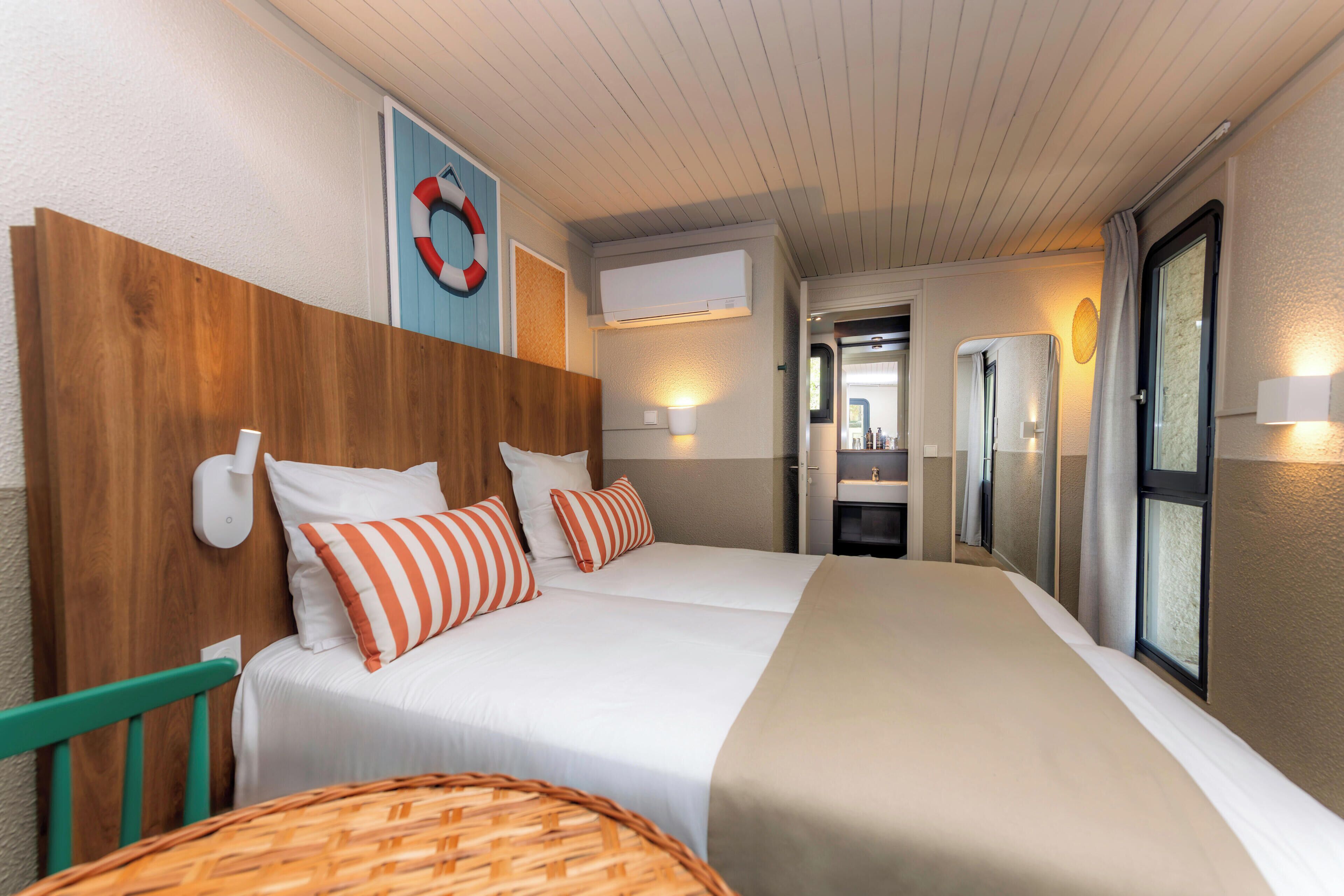 Ein Hotelzimmer mit zwei Betten, einem hölzernen Kopfteil, einer wandmontierten Klimaanlage, einem durch eine Glastür einsehbaren Badezimmer und einem Korbstuhl.