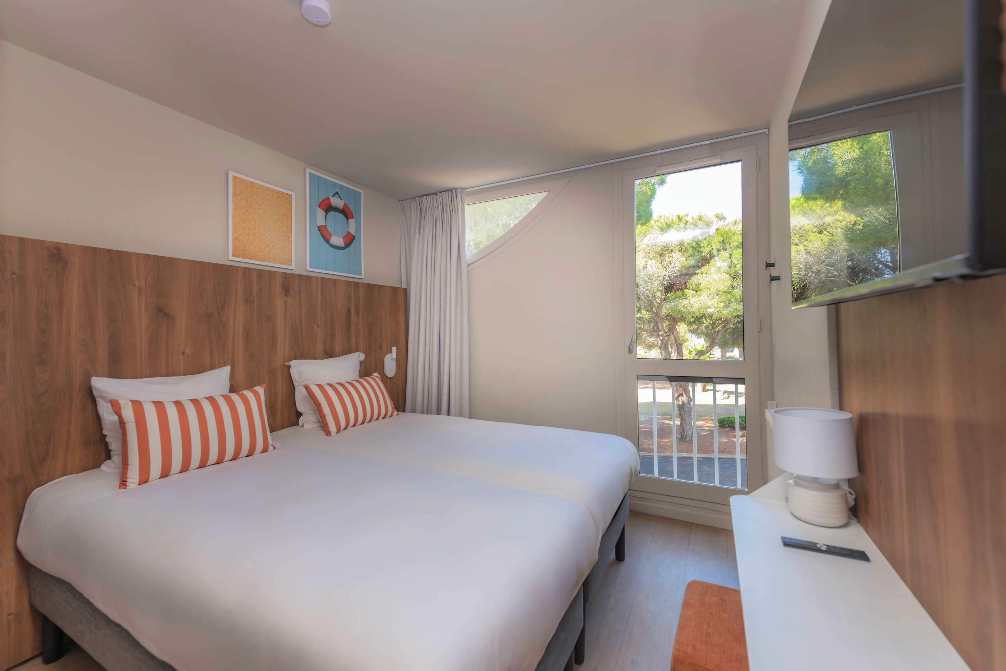 A hotel room with a bed, a bedside table with a lamp, a window with a view of trees, and a wall-mounted TV.