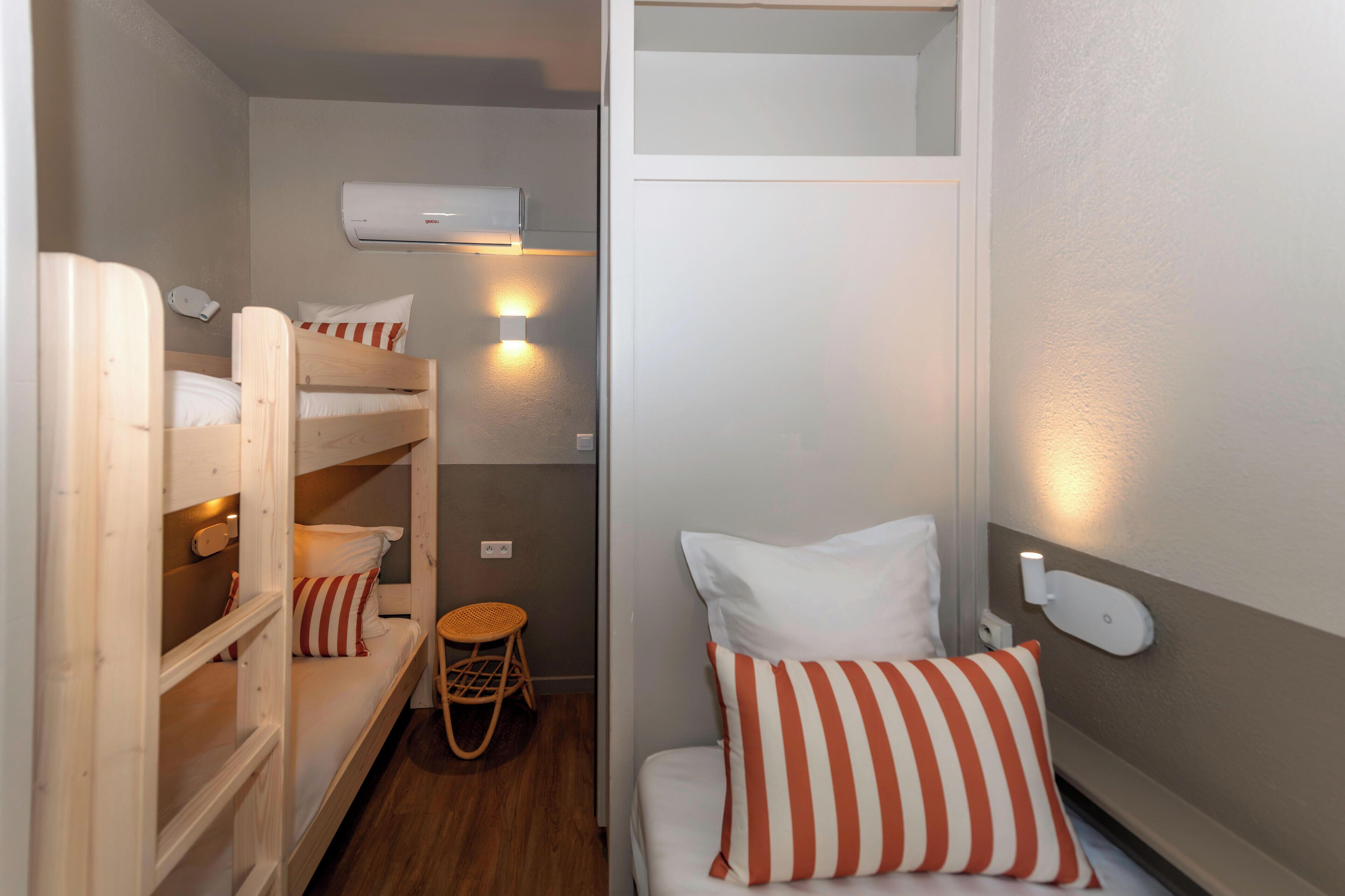 Une petite pièce avec des lits superposés, un petit tabouret et un luminaire fixé au mur.