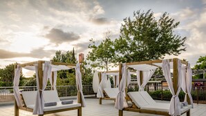 Outdoor wooden canopy beds with white cushions, arranged on a tiled patio with a view of trees and a cloudy sky.