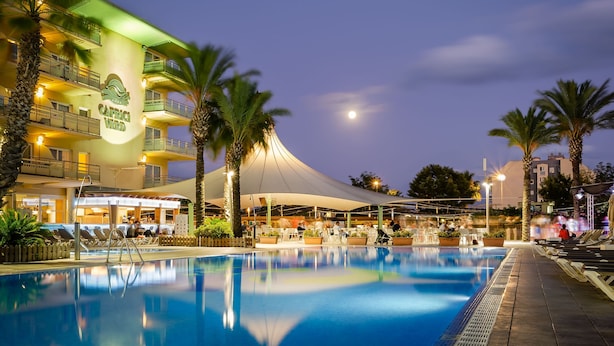 A hotel with a swimming pool, palm trees, and a covered area with people.