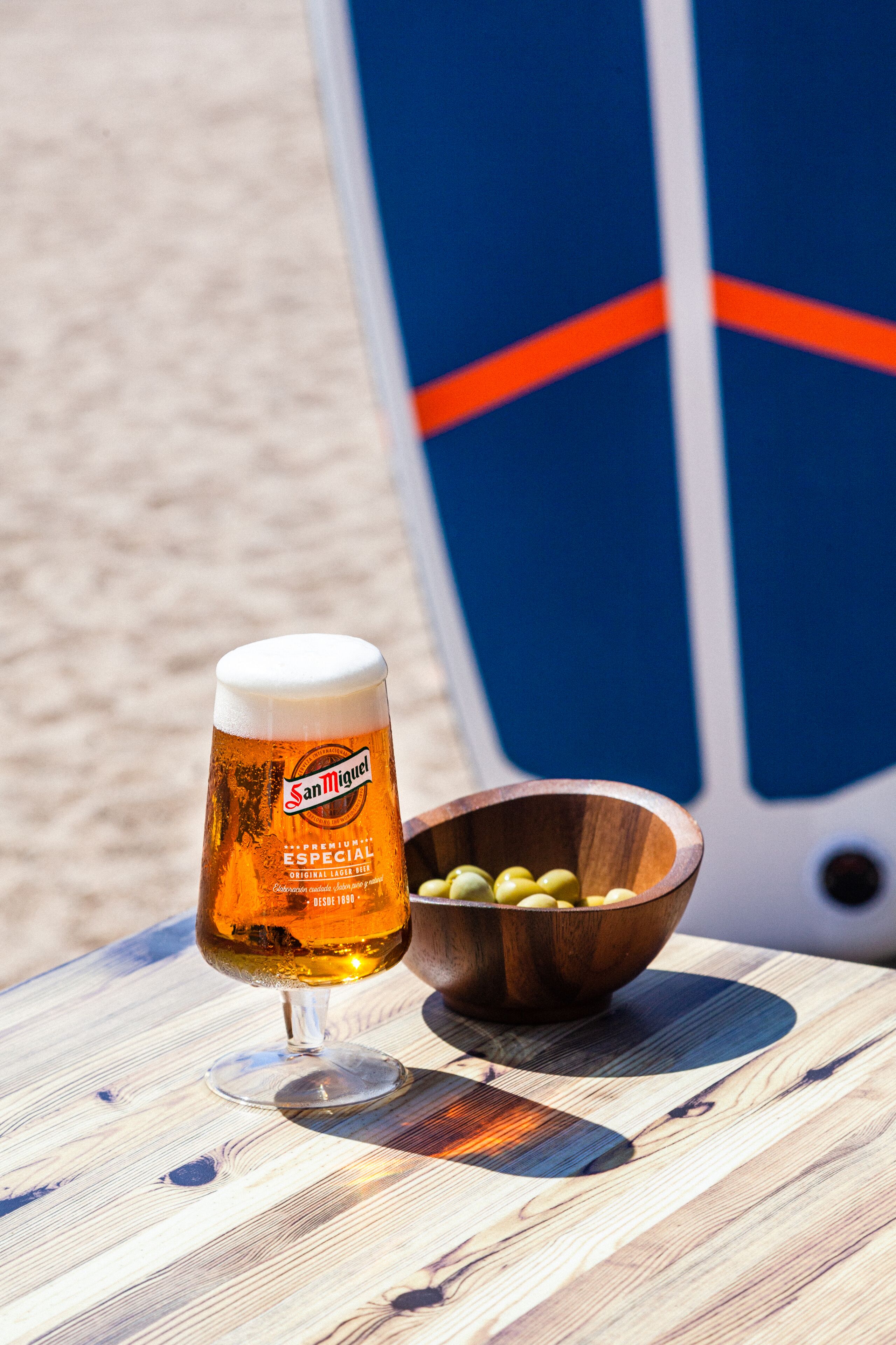 Een glas San Miguel bier, een houten schaal met olijven en een surfboard op de achtergrond.