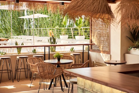 An outdoor seating area with wicker chairs and tables, potted plants, and a view of greenery through a fence.