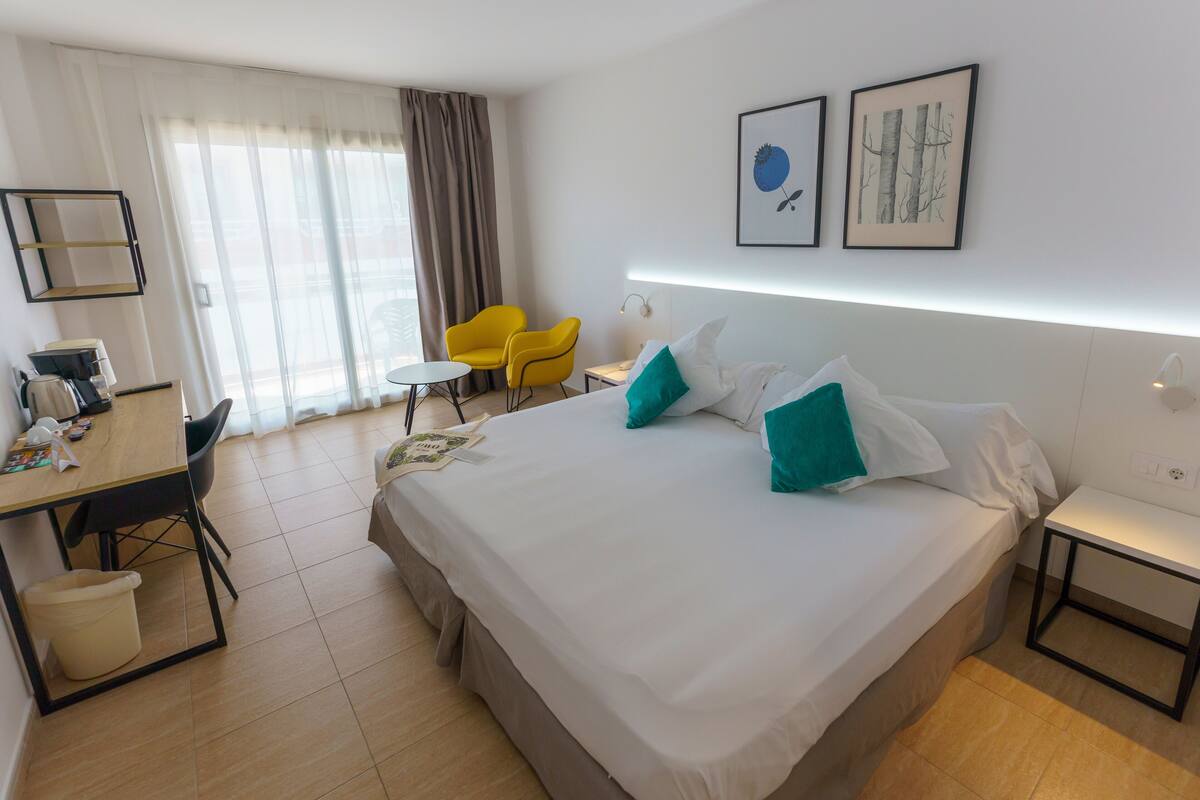A hotel room with a large bed, a desk with a chair, a small table, and a trash bin. There are two yellow chairs and a window with curtains.