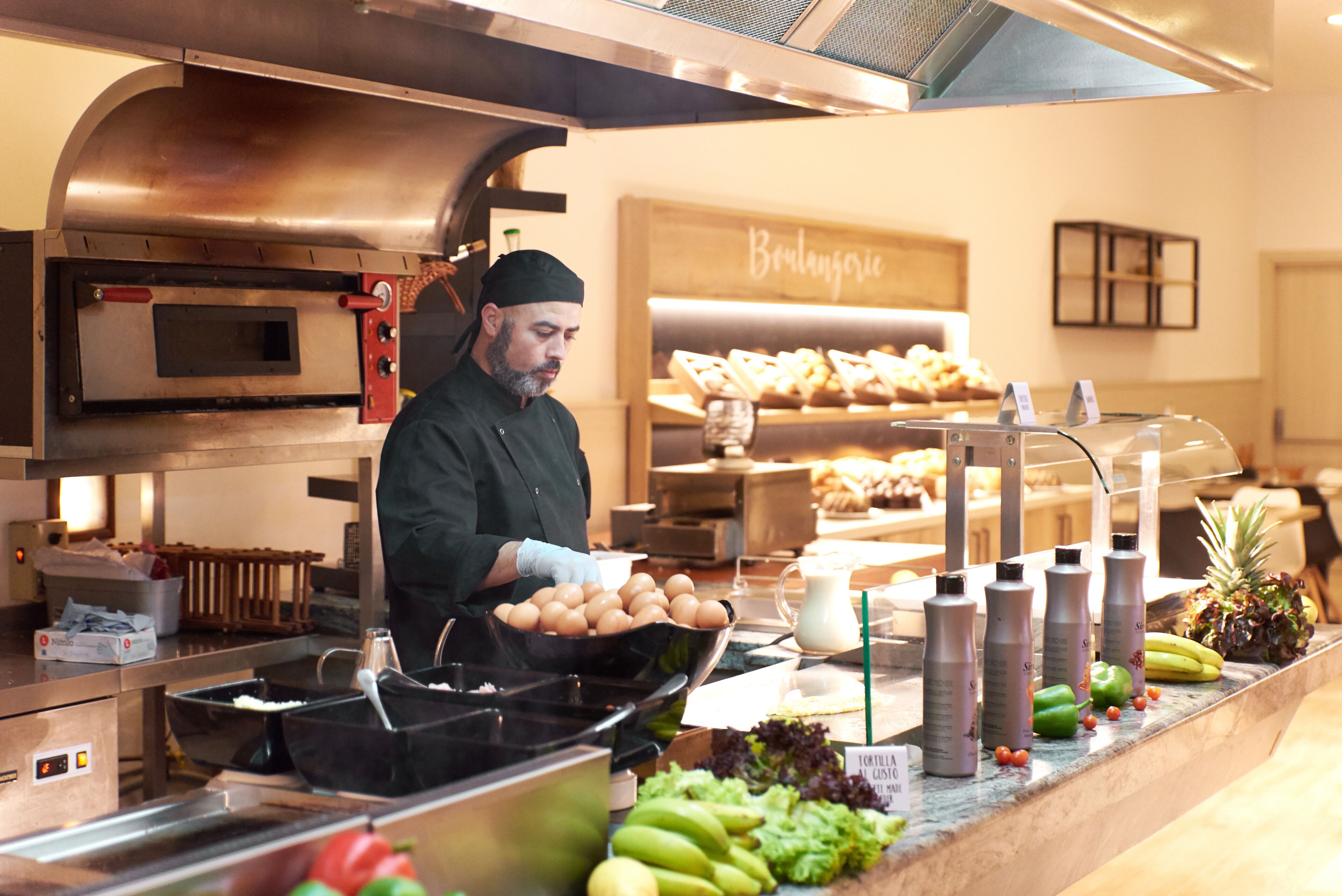 Een kok bereidt eten in een keuken met een roestvrijstalen oven en een presentatie van gebak op de achtergrond.